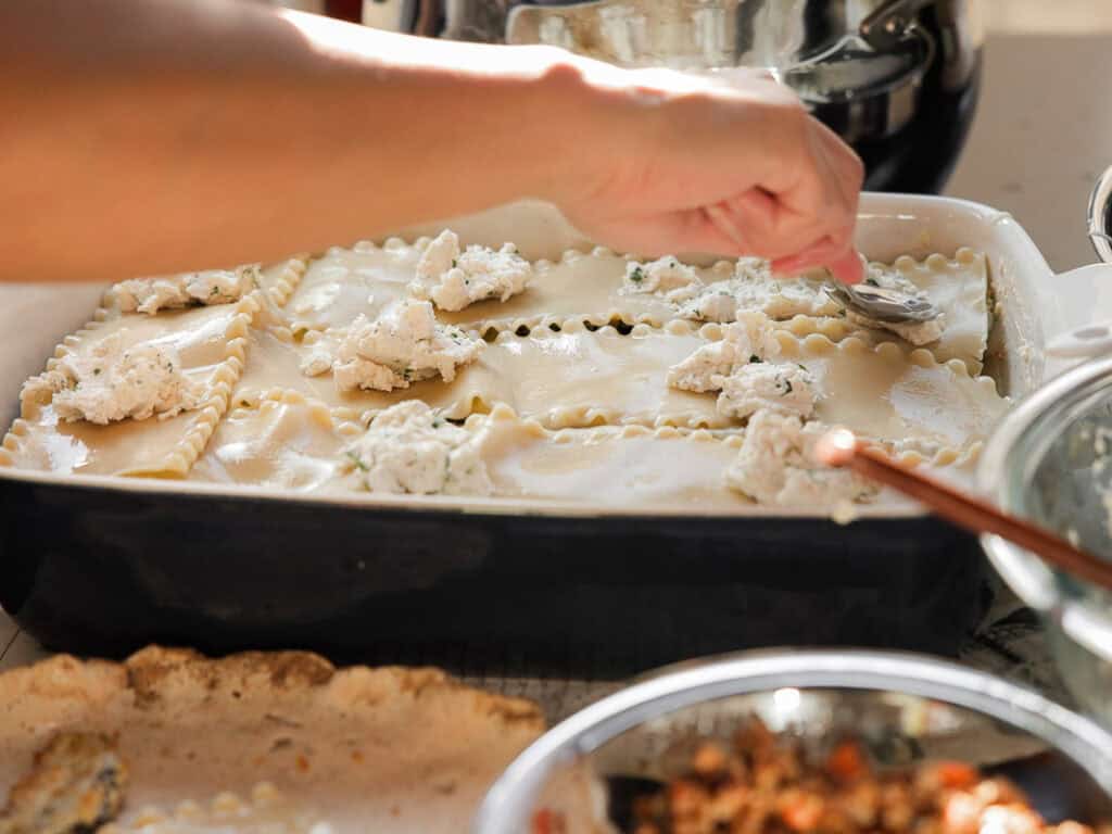 A close-up of a person assembling vegan lasagna in a baking dish. They are spreading ricotta cheese on layers of pasta sheets with a spoon. Nearby, there are ingredients and kitchen utensils on the countertop.